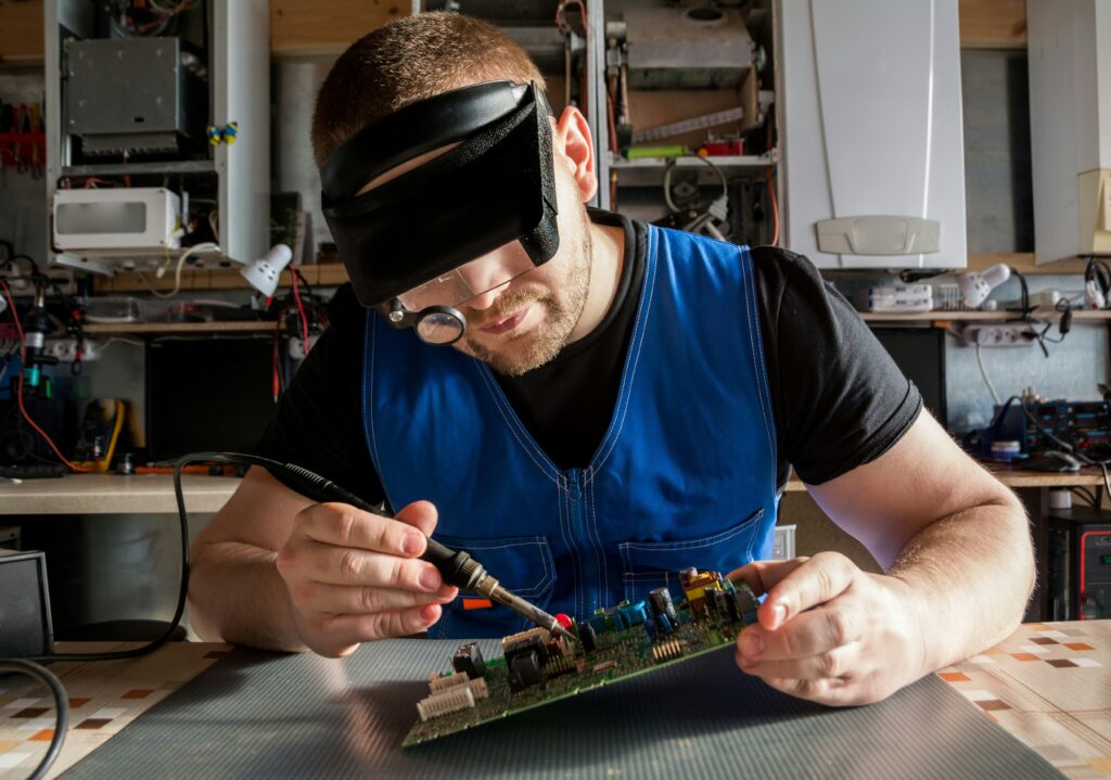 A person wearing a magnifying headset and blindfold-style visor closely soldering a circuit board