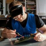 A person wearing a magnifying headset and blindfold-style visor closely soldering a circuit board